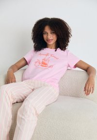 T-shirt de algodão rosa com gráfico "Costa Amalfitana" em laranja. Combinado com calças de pijama listradas em creme suaves. Sofá de tecido texturizado branco e aconchegante.