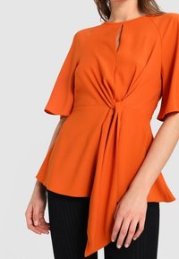 Blouse péplum orange avec détail noué à la taille, manches courtes, encolure ronde et texture de tissu lisse. Associée à un pantalon à fines rayures.