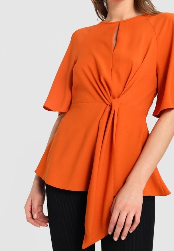 Blouse péplum orange avec détail noué à la taille, manches courtes, encolure ronde et texture de tissu lisse. Associée à un pantalon à fines rayures.