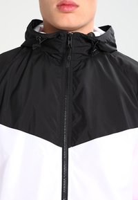 Veste noir et blanc avec fermeture éclair sur le devant, capuche et design en blocs de couleur contrastants. Fabriquée en tissu lisse et léger. Présente un cordon de serrage pour un ajustement.