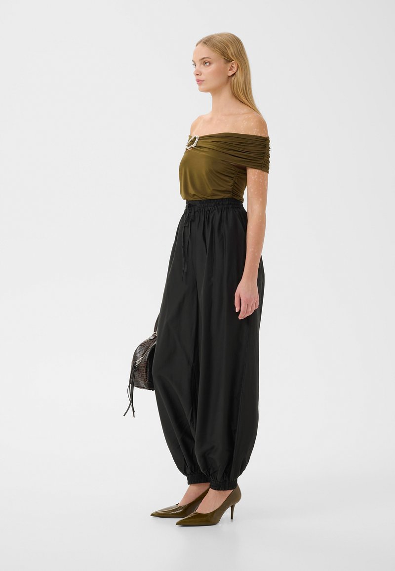 Femme portant un top olive épaules dénudées avec broche, pantalon noir ample taille haute, talons olive, tenant un petit sac à main, tournée vers la droite.