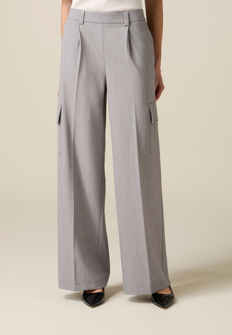 Mujer vistiendo pantalones anchos de pierna ancha en color gris claro con plisados y bolsillos laterales tipo cargo, combinados con zapatos negros de punta afilada.