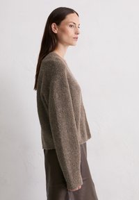 Brązowy, teksturowany sweter z długim rękawem, charakteryzujący się luźnym krojem i okrągłym dekoltem, noszony z ciemną spódnicą. Minimalistyczny design, brak widocznych wzorów.