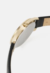 Reloj en tono dorado con una caja redonda, correa de cuero negra y una corona texturizada. Parte trasera suave en tono plateado con un diseño minimalista.