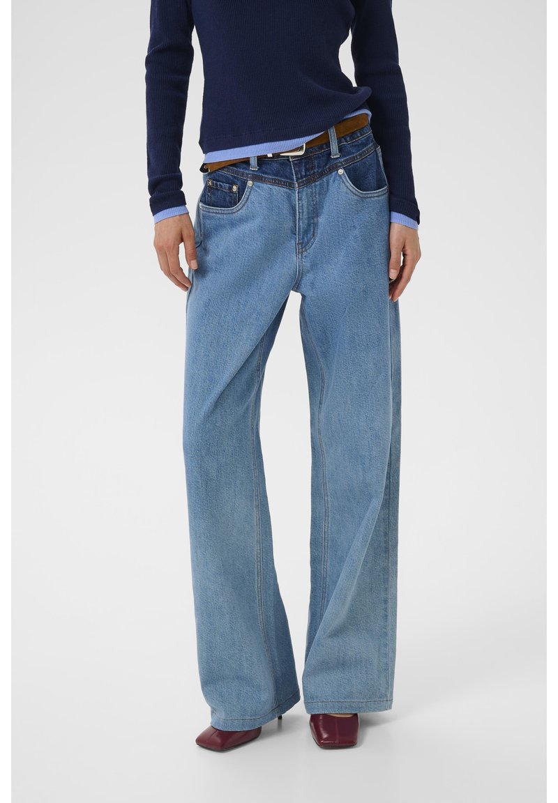 Personne portant un jean bleu bicolore à jambes larges avec une ceinture marron, un pull bleu marine et des chaussures à talons bordeaux sur fond blanc.