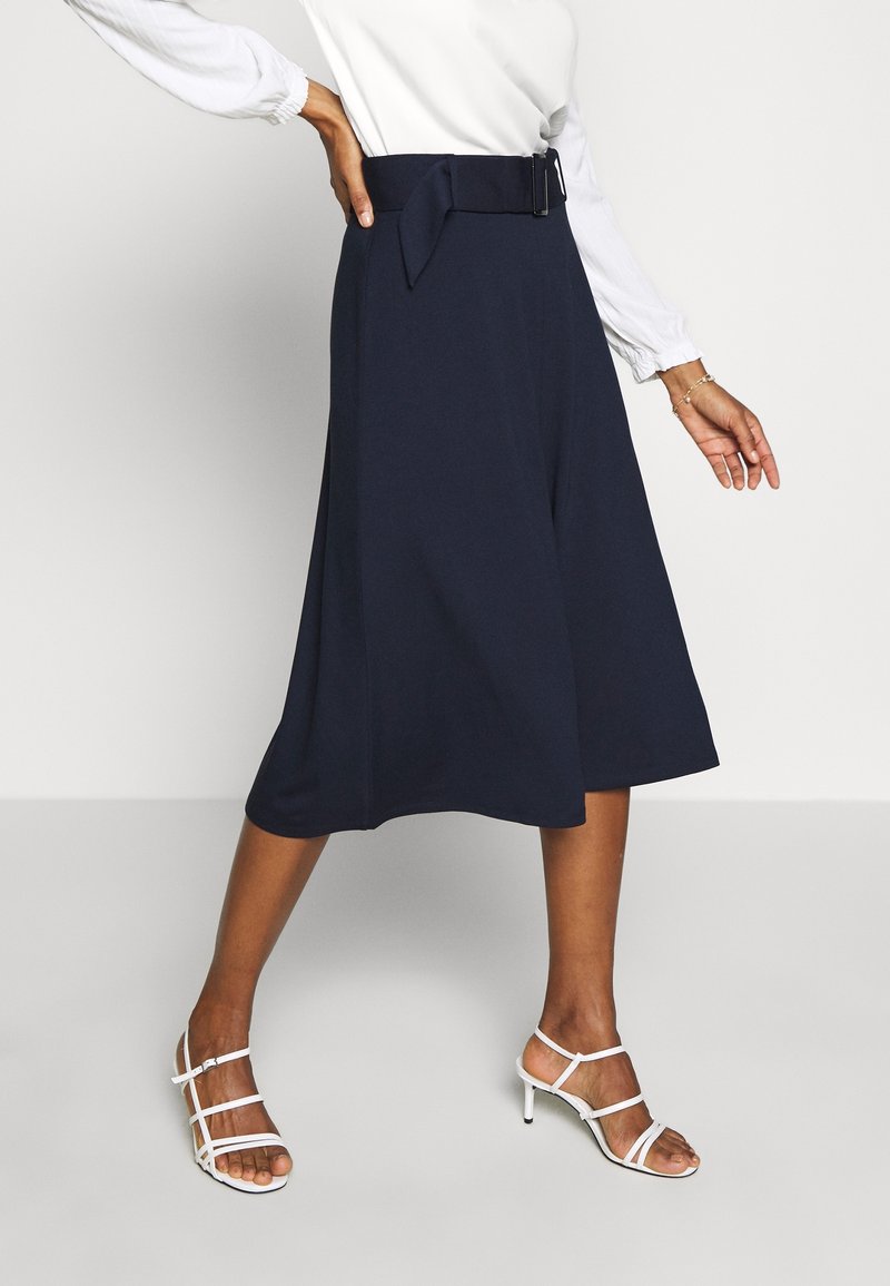 Femme portant une jupe midi bleu marine avec ceinture, un chemisier blanc à manches longues et des sandales blanches à talons et brides, debout sur un sol gris.