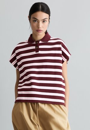 Jeune femme portant un polo à manches courtes rayé bordeaux et blanc, et un pantalon beige, se tenant devant un fond uni gris clair.