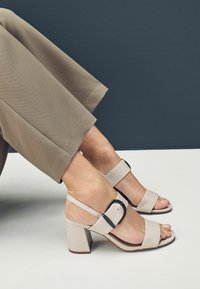 Beige blockhælede sandaler med brede stropper og en sort spænde detalje, der viser en glat tekstur. Båret med beskåret beige bukser.