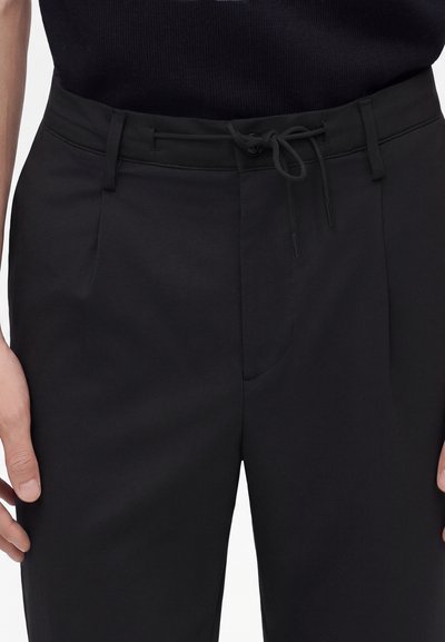 Gros plan d'une personne portant un pantalon noir avec un cordon à la taille et des passants de ceinture au-dessus d'un haut noir, les mains reposant sur les côtés.