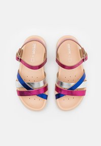 Sandale avec des brides métalliques multicolores en rose, bleu et argent, dotée d'une semelle en beige clair et d'une boucle de cheville réglable.