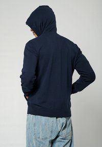 Hoodie marine avec un ourlet côtelé et des patchs aux coudes, doté d'une fermeture éclair à l'avant et d'un cordon de serrage réglable, fabriqué à partir d'un tissu doux.