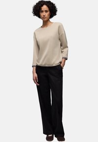 Frau mit lockigem Haar, die ein beigefarbenes langärmliges Oberteil, schwarze weite Hosen und braune Schuhe trägt, mit der Hand in der Tasche vor weißem Hintergrund steht.