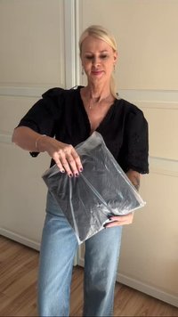 Mujer con cabello rubio vestida con blusa negra y jeans azules sostiene un paquete de plástico sellado que contiene un objeto negro doblado en interiores.
