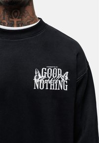 Felpa nera con una grafica stampata bianca che riporta il testo "Presentato da Good For Nothing", abbellita con design di farfalle.