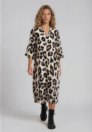 Frau mit welligem Haar, die ein knieumspielendes beiges und schwarzes Kleid mit Leopardenmuster und schwarze Schnürstiefel vor einem einfarbigen Hintergrund trägt.