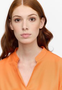 Blusa arancione brillante con colletto alla mandarino, texture liscia e lucentezza sottile; presenta una vestibilità morbida e linee pulite.