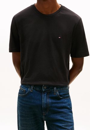 Mann trägt ein einfaches schwarzes T-Shirt mit kurzen Ärmeln und kleinem Logo auf der Brust sowie blaue Jeans und steht mit den Händen hinter dem Rücken.