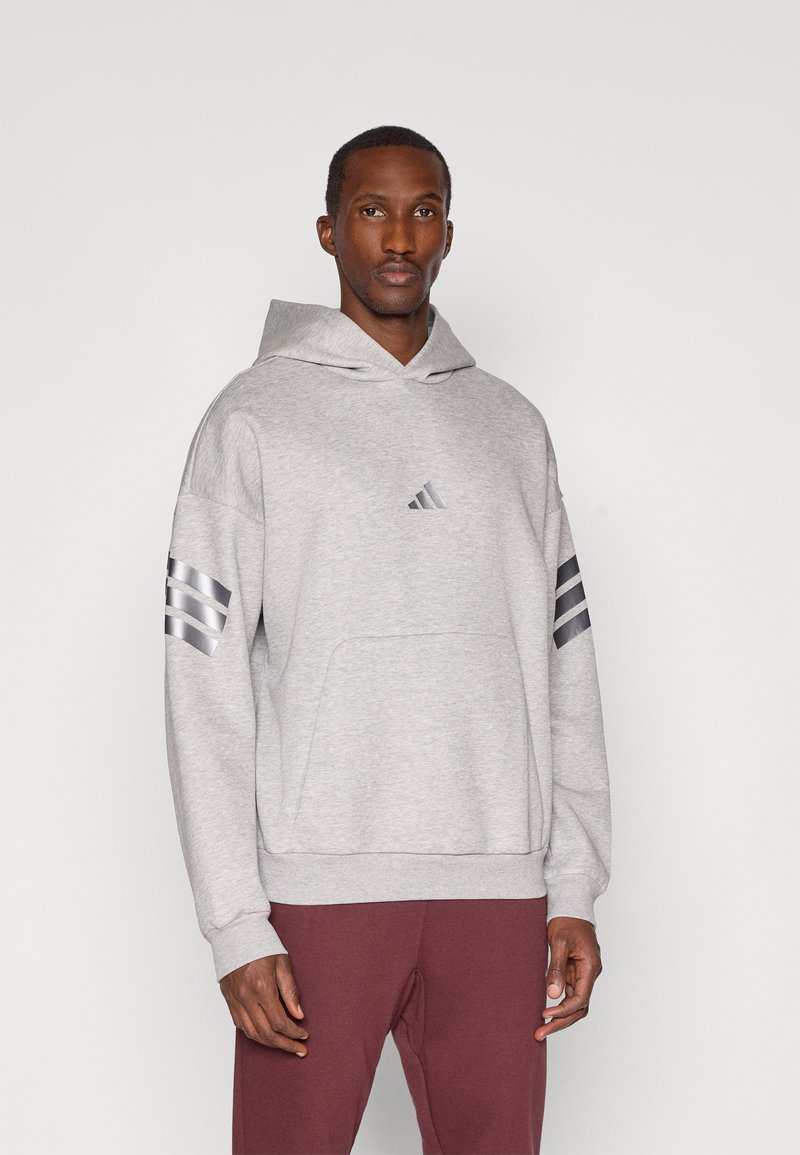 Homem a usar um hoodie cinza claro com riscas pretas nas mangas e um logótipo no peito, combinado com calças bordô, de pé contra um fundo branco.