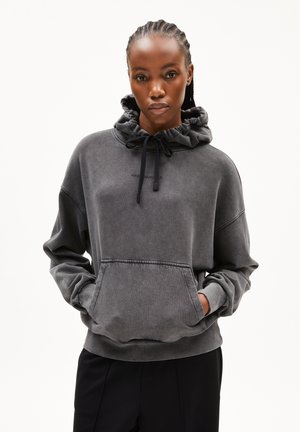 Vrouw met gevlochten haar, gekleed in een vervaagde zwarte hoodie met een gekreukte kraag en een kangoeroezak, met haar handen erin gestoken, tegen een effen achtergrond.