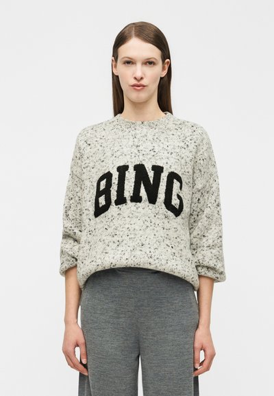 Femme aux cheveux bruns et raides portant un pull gris moucheté avec le mot « BING » en lettres noires et un pantalon gris ample, debout devant un fond blanc.