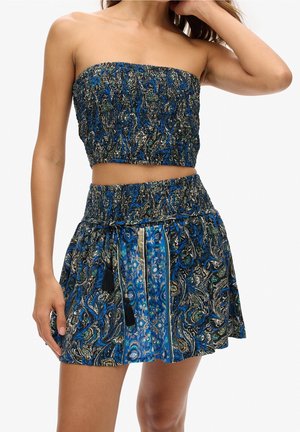 Femme portant un top court bleu et noir à smocks sans bretelles et une jupe taille haute assortie avec des cordons à pompons.