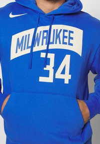 Modrá mikina Nike s velkým bílým logem "MILWAUKEE", číslem "34", klokankovou kapsou a tkánkami na kapuci.