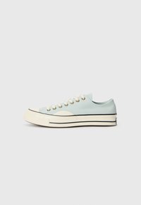 Light blue canvas sneaker with a rounded toe, white rubber sole, and black stripes. Features gold eyelets and flat laces.