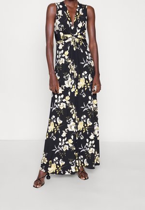 Robe longue noire sans manches à imprimé floral jaune et blanc, col en V plongeant, taille froncée, portée avec des sandales à talons noires à brides.