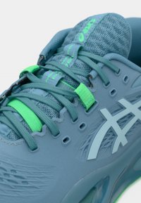 Αθλητικό παπούτσι ανοιχτού μπλε χρώματος με επάνω μέρος από δίχτυ, πράσινες λεπτομέρειες, επίπεδοι κορδόνια και εμφανές λογότυπο. Υπόδημα με υφή, αυλακώσεις και οπές αερισμού.