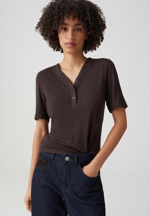 Junge Person mit lockigem Haar, die ein dunkelbraunes kurzärmeliges Hemd mit Knöpfen und dunkelblaue Jeans trägt, mit einer Hand in der Hosentasche.