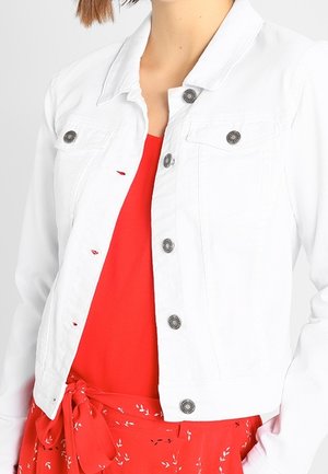 Femme portant une veste blanche boutonnée sur une robe rouge avec une taille nouée et de petits motifs floraux blancs.
