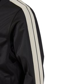 Svart jakke med en teksturert finish, med en kremfarget og svart stripet detalj langs skulderen. Slim fit-design med et sporty utseende.