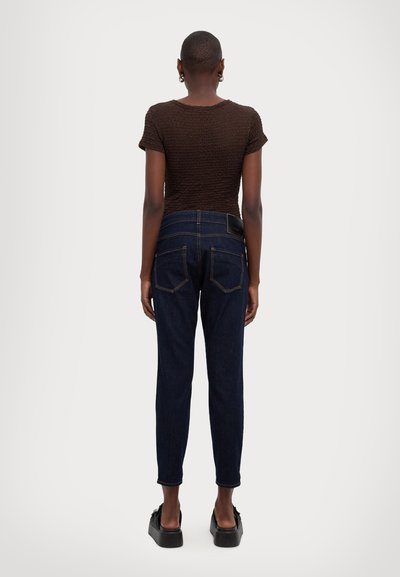 Σκούρο μπλε τζιν παντελόνι ψηλής μέσης με εφαρμοστή σιλουέτα, αντίθετες ραφές και δύο πίσω τσέπες. Το μπλουζάκι είναι καφέ με υφή.