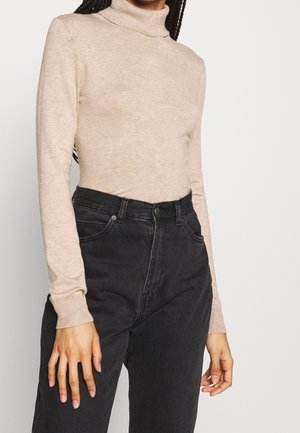 Kvinde iført en beige rullekravetrøje stoppet ned i sorte, højtaljede jeans, stående mod en neutral baggrund.