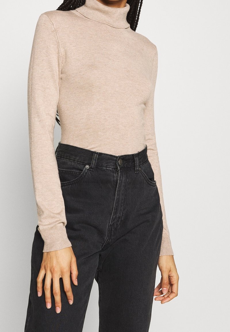 Femme portant un pull à col roulé beige rentré dans un jean noir taille haute, debout devant un fond neutre.