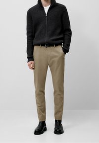 Donkergrijze geribbelde ritstrui, tan rechte broek en zwarte leren laarzen. Opvallende details zijn een hoge kraag en gladde texturen.