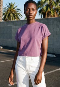 Joven mujer con una camiseta corta morada y pantalones blancos de talle alto, de pie al aire libre cerca de una pared con palmeras.