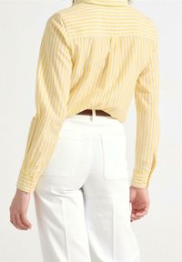 Gul- og hvidstribet bluse med krave og knapærmer, parret med hvide højderede bukser. Bagsiden viser designdetaljer.