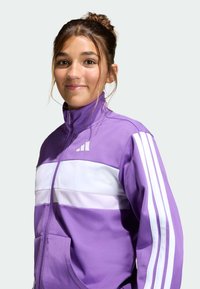 Violetti takki, jossa valkoiset raidat rinnalla ja hihoissa, vetoketjullinen kaulus, pehmeä materiaali ja logo vasemmalla yläpuolella.