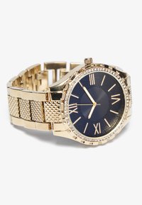 Montre dorée avec un bracelet texturé, un cadran bleu, des index en chiffres romains et une lunette ornée de petits cristaux.