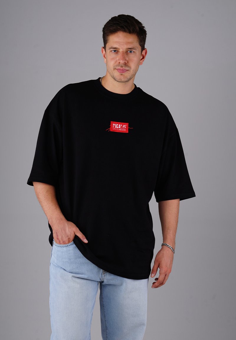 Mann in einem schwarzen, übergroßen T-Shirt mit rotem Logo und hellblauen Jeans, der vor einem einfarbigen grauen Hintergrund steht.