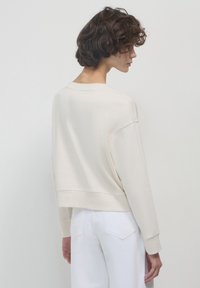 Felpa beige chiaro con un design cropped, maniche lunghe, polsini a costine e un ampio scollo a giro, mostrata da dietro.