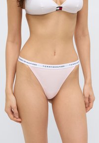 Φως ροζ στρινγκ με λευκή ζώνη μέσης που φέρει το λογότυπο "TOMMY HILFIGER". Το υλικό φαίνεται λείο και ελαστικό, σχεδιασμένο για άνεση.