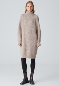 Strickpulloverkleid in hellbeige mit strukturiertem Finish, langen Ärmeln und seitlichen Schlitzen, kombiniert mit schwarzen Strumpfhosen und braunen Absatzstiefeln.