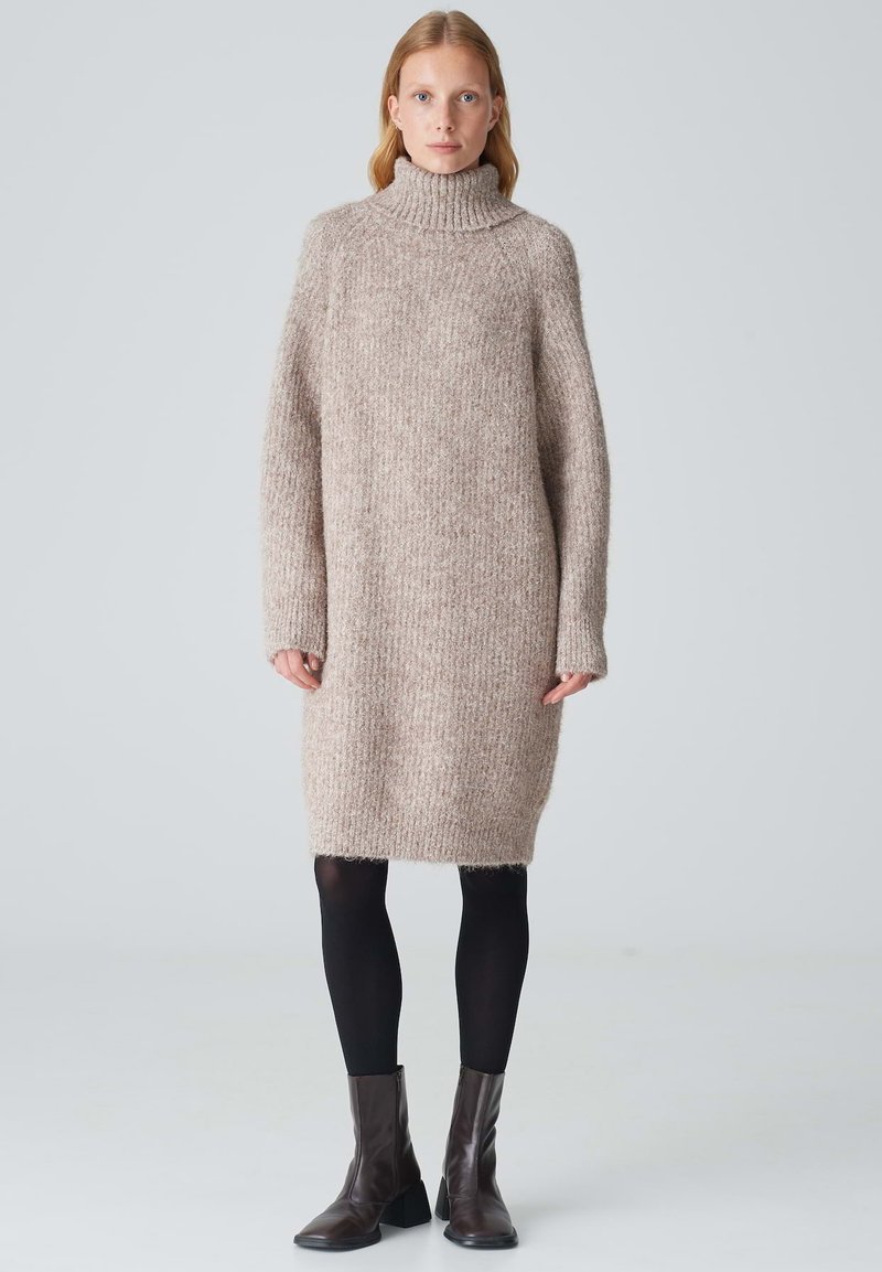 Strickpulloverkleid in hellbeige mit strukturiertem Finish, langen Ärmeln und seitlichen Schlitzen, kombiniert mit schwarzen Strumpfhosen und braunen Absatzstiefeln.