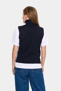 Mornarska pletena brezrokavna turtlenecka, z rebrastim robom, nošena čez belo majico, v paru z ohlapnimi denim hlačami.