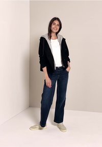 Svart och grå huvtröja över en vit skjorta, tillsammans med blå vida jeans och ljusa sneakers, mot en neutral bakgrund.