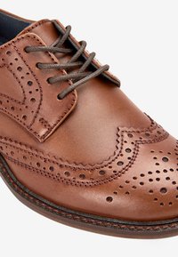 Brown leather dress shoe with brogue detailing, featuring perforations, laces, and a contrasting dark sole. Smooth finish and structured design.