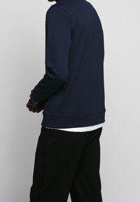 Man i marinblå långärmad skjorta över vit t-shirt och svarta byxor, som står mot en enkel ljusgr å bakgrund.