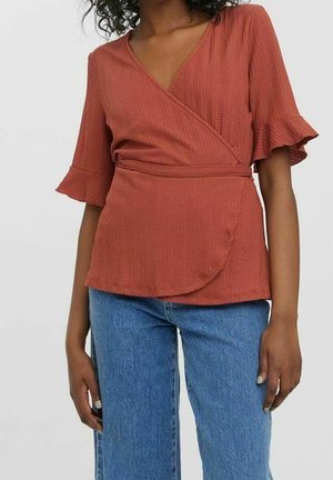 Femme portant un haut cache-cœur couleur rouille avec des manches courtes à volants et un jean bleu, debout devant un fond clair uni.